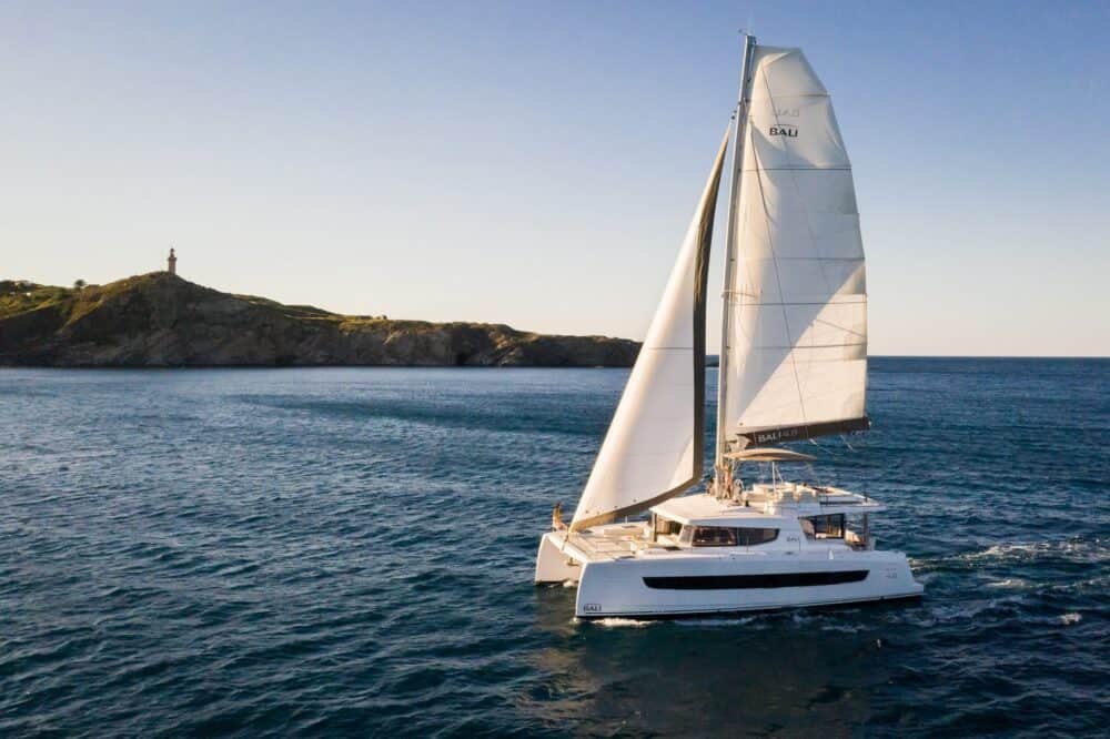 Bali 4.8 catamaran charter Marmaris under full main and self-tacking jib passing a rocky headland at golden hour