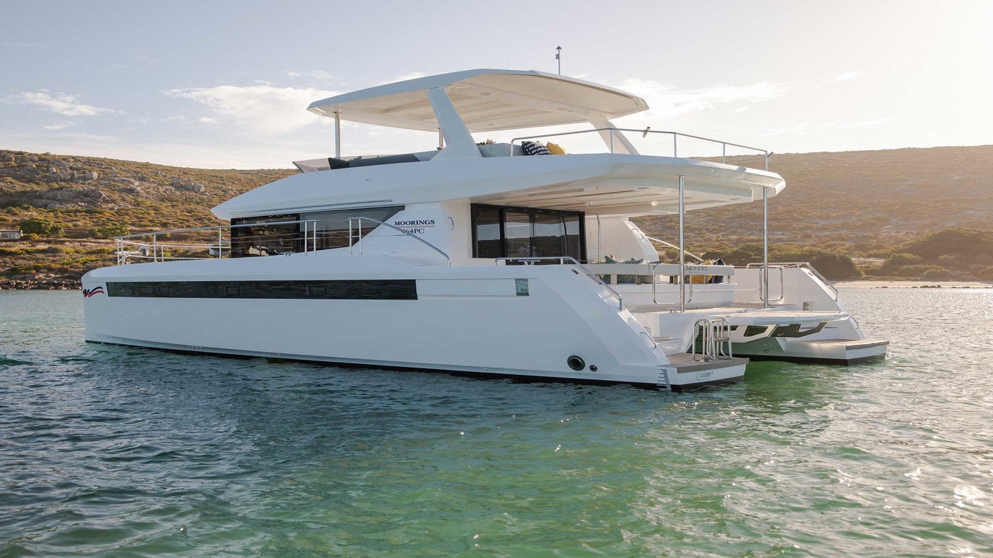 Moorings 464PC catamaran at anchor viewed from the stern quarter, flybridge bimini extended over the upper helm