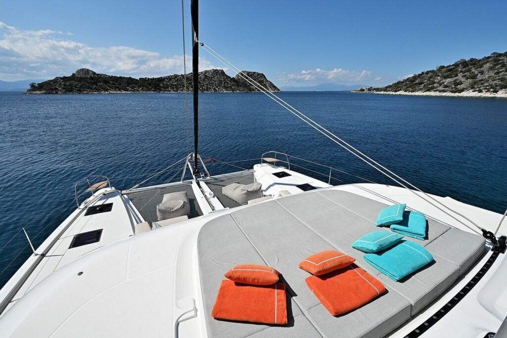 Lagoon 55 catamaran charter Kos - Lagoon 55 catamaran foredeck lounge sunbeds with guests relaxing, anchored beneath limestone cliffs, Kos charter UT1704